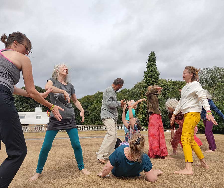 Un grupo de personas baila y se mueve alegremente en un campo de hierba con árboles y un cielo nublado de fondo. Los participantes llevan ropa informal y colorida y parecen estar participando en una actividad lúdica.
