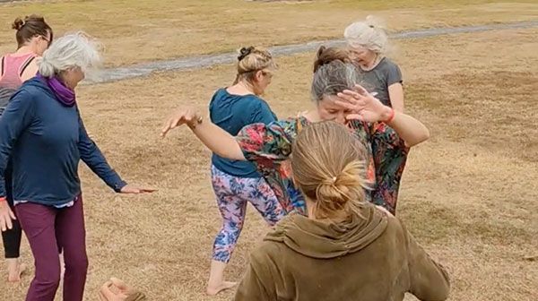 Un grupo de personas baila alegremente al aire libre en un campo de hierba. Llevan ropa informal y parecen participar en una sesión de baile libre y expresivo.