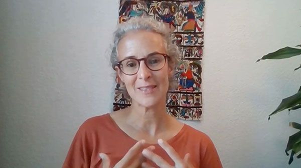Una persona con cabello gris rizado y gafas sonríe. Lleva una camiseta naranja y hace gestos con las manos. En la pared detrás de ella hay una obra de arte colorida y a la derecha se ve parcialmente una planta.